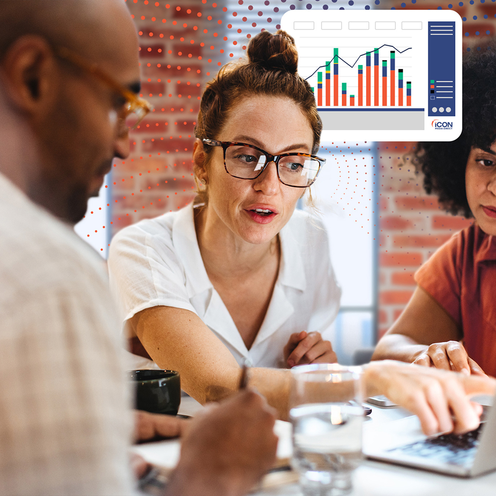 Woman shows colleagues laptop data; graph shows upward trend; office setting.
